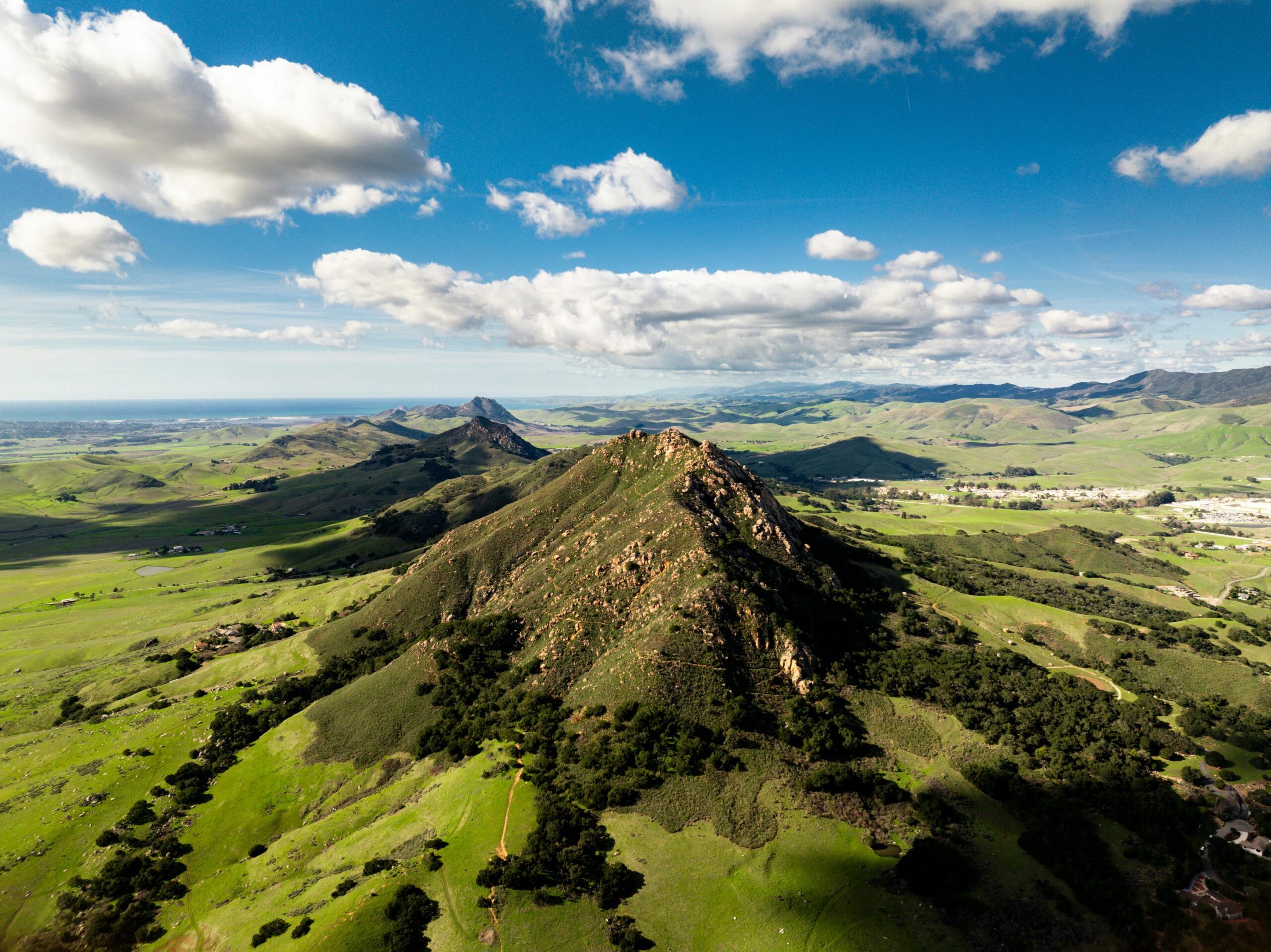 Aerial view of green rolling hills and volcanic peaks near San Luis Obispo under a blue sky with clouds